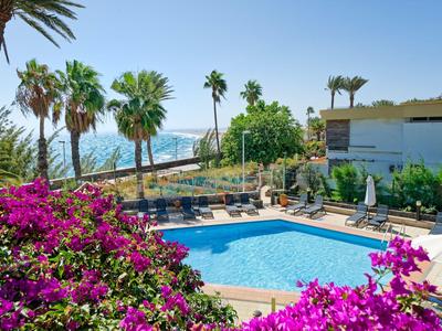 Espace piscine avec bougainvilliers en fleurs, palmiers et vue sur la mer à côté d'un bâtiment hôtelier.