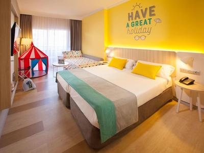 Modernes Hotelzimmer mit Doppelbett, Kinderbett, buntem Zelt und gelber Wand mit Schriftzug.