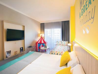 Helles Hotelzimmer mit großem Bett, buntem Kinderspitzzelt und gelber Akzentwand.