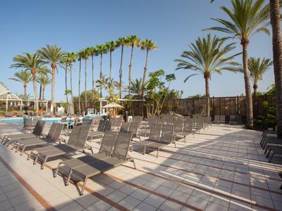 Leere Liegestühle am Pool mit Palmen und Sonnenschirmen unter blauem Himmel.