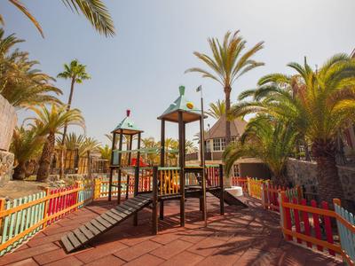 Bunter Spielplatz mit Klettergerüst, Rutschbahn und Palmen bei sonnigem Himmel.