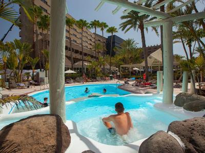 Hotelpool mit Palmen, Liegestühlen und Gästen, umgeben von mehrstöckigem Gebäude.