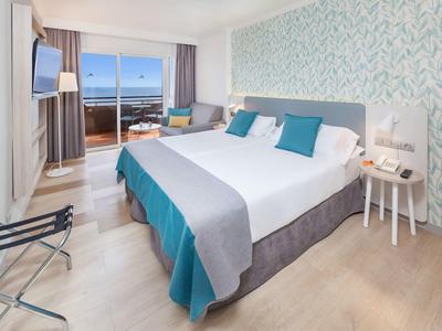 Helles Hotelzimmer mit großem Bett, türkisfarbenen Akzenten und Balkon mit Meerblick.