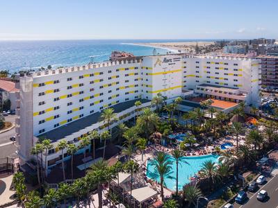 Large hotel with pool and palm trees by the sea under clear sky.