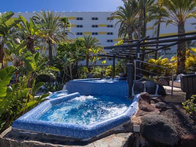 Hotel area with a jacuzzi surrounded by palm trees and a multi-story building in the background.