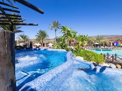 Outdoor pool with blue mosaics, palm trees, and sun umbrellas under clear sky.