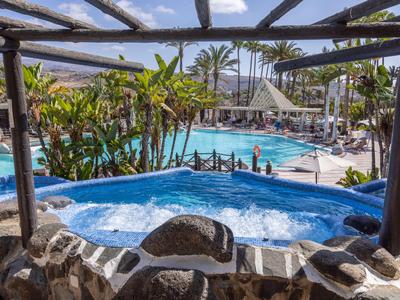 Hot tub overlooking an outdoor pool surrounded by palm trees and sun umbrellas.