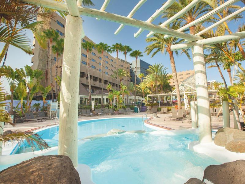 Espace piscine d'hôtel avec palmiers, bains à remous et terrasse ensoleillée sous ciel bleu.