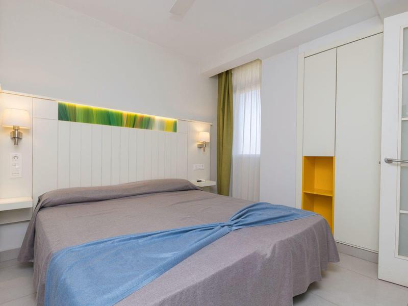 Jasny pokój hotelowy z podwójnym łóżkiem, dwoma stolikami nocnymi i wbudowaną szafą z żółtą półką.