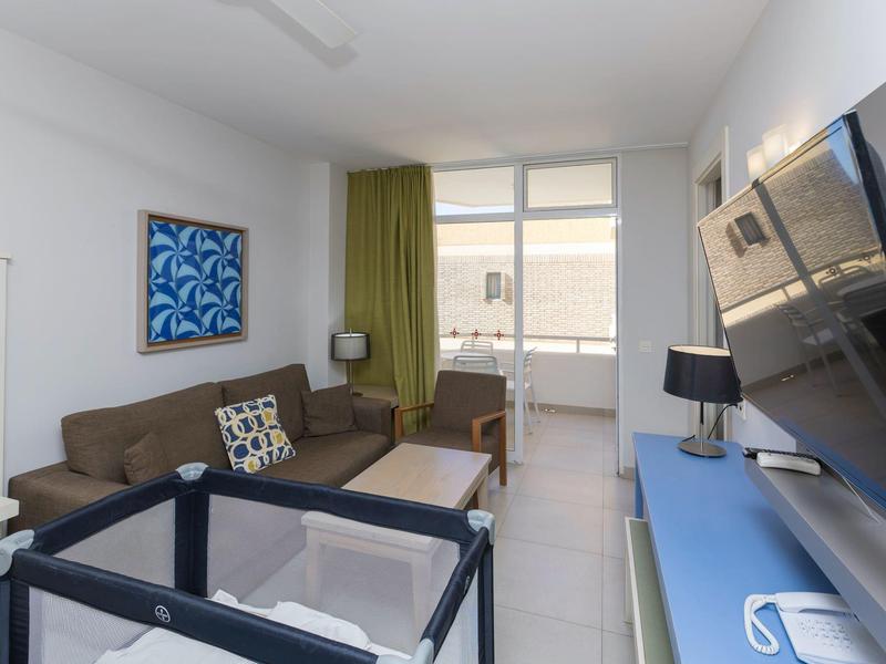 Nowoczesny pokój hotelowy z sofą, łóżeczkiem dziecięcym, telewizorem i balkonem z widokiem na zewnątrz.