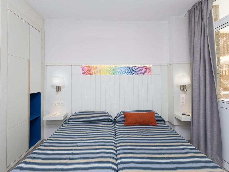 Dwa pojedyncze łóżka z pasiastymi narzutami w jasnym pokoju hotelowym z oknem i zasłoną.