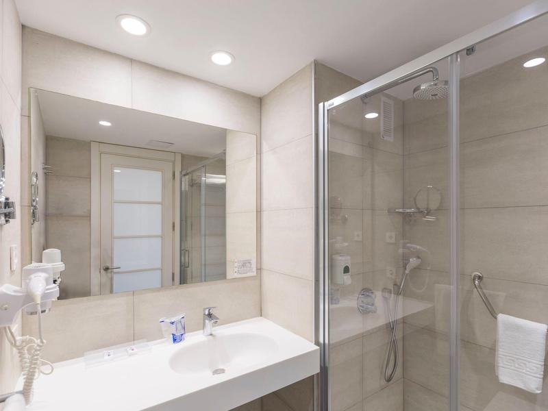Nowoczesna łazienka hotelowa z dużym prysznicem, umywalką i neutralnymi płytkami.