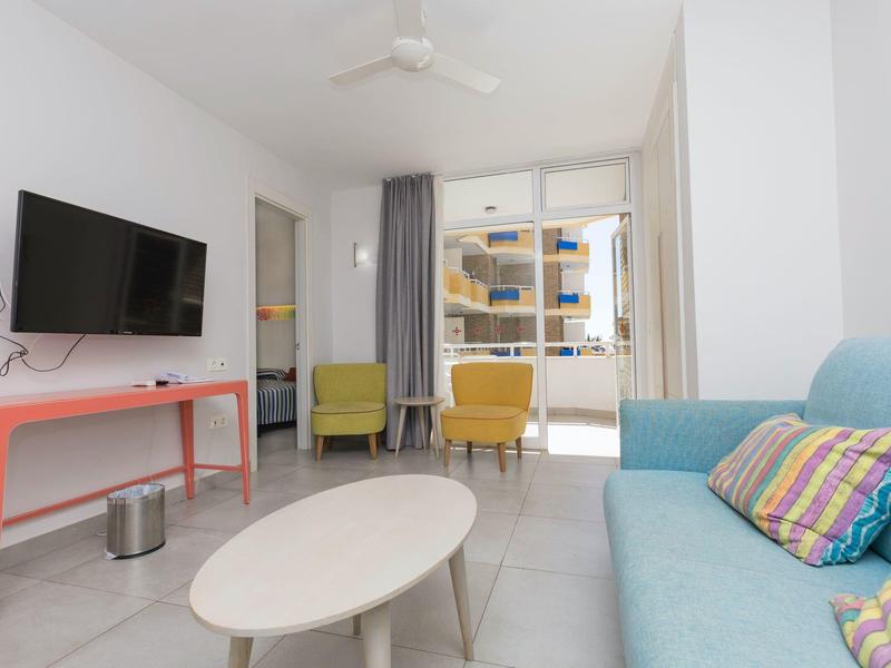 Jasny salon z sofą, telewizorem, fotelem i balkonem z widokiem na budynki mieszkalne.