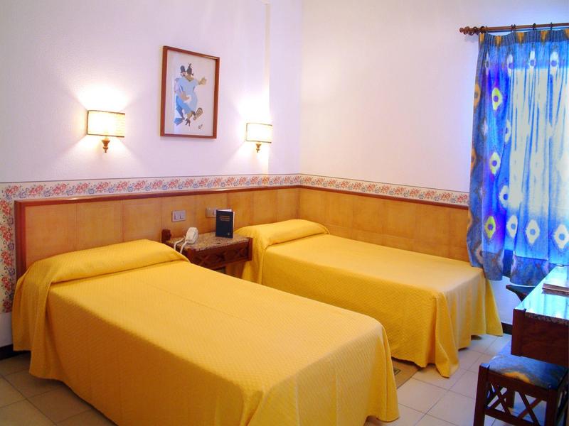 Pokój hotelowy z dwoma pojedynczymi łóżkami, żółtymi narzutami i oświetlonym stolikiem nocnym.