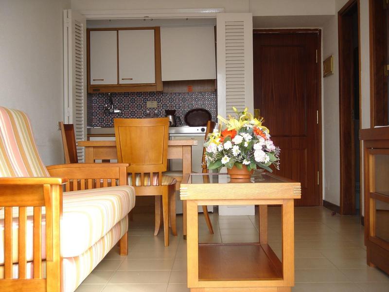 Mały salon z sofą, drewnianym stołem, krzesłami i otwartą aneksem kuchennym w tle.