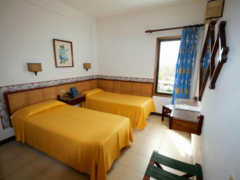 Pokój hotelowy z dwoma pojedynczymi łóżkami z żółtymi narzutami i oknem z niebieskimi zasłonami.