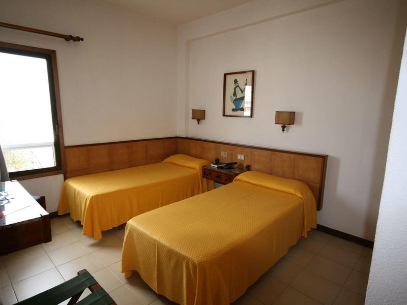 Prosty pokój hotelowy z dwoma łóżkami pojedynczymi i żółtymi narzutami, okno z zasłoną.