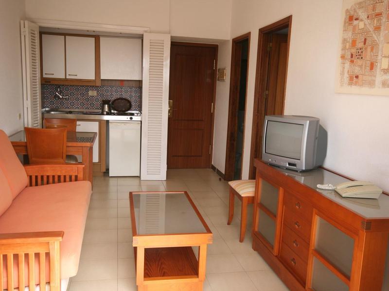 Mały pokój dzienny z drewnianymi meblami, kanapą, szklanym stołem i aneksem kuchennym w pokoju hotelowym.