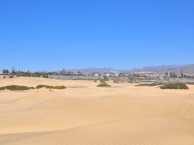 Weite Sanddünenlandschaft vor einer Stadt unter klarem blauen Himmel.