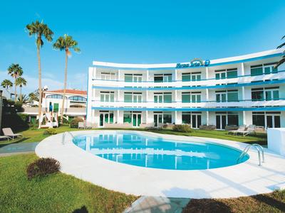 Modernes, weißes Hotel mit blauem Pool, Palmen und grünem Rasen unter blauem Himmel.
