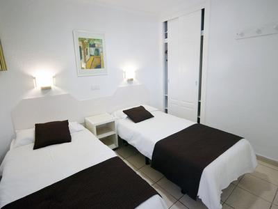 Einfaches Zimmer mit zwei Einzelbetten, weißen Decken, dunklen Kissen und Wandlampen.