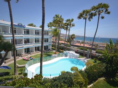Hotel mit rundem Pool, Palmen, Liegestühlen und Meer im Hintergrund bei sonnigem Himmel.