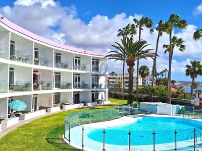 Modern hotel met balkons, rond zwembad, palmbomen en zeezicht onder een blauwe lucht.