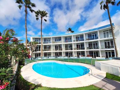 Een modern hotel met een ronde pool, omringd door palmbomen en bloeiende planten onder een blauwe lucht.