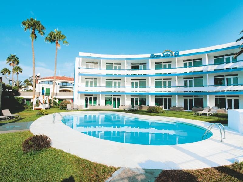 Moderner Hotellkomplex mit rundem Pool und Palmen unter klarem blauem Himmel.
