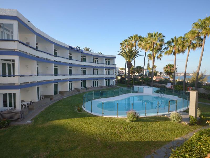 Modernes Hotel mit weißer Fassade, Swimmingpool und hohen Palmen bei klarem Himmel.