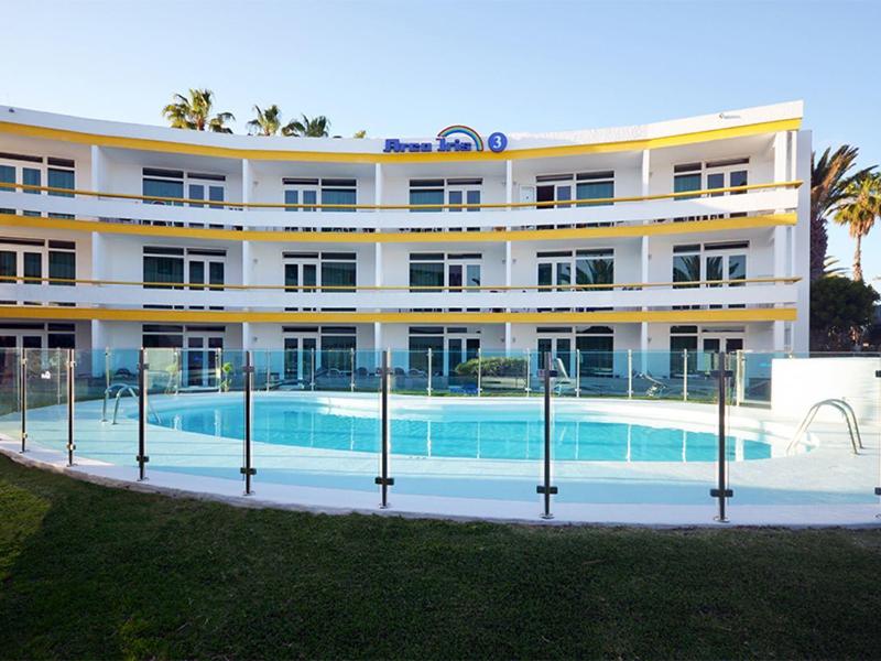 Modernes Hotelgebäude mit Balkonen rund um einen runden, umzäunten Außenpool unter blauem Himmel.