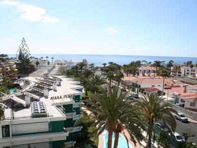 Blick auf Küstenstadt mit Palmen, Dächern weißer Gebäude und blauem Meer im Hintergrund.