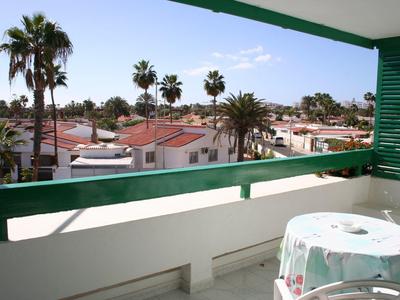 Balkon mit grünem Geländer, Tisch mit Tuch, Palmen und weiße Häuser unter blauem Himmel.