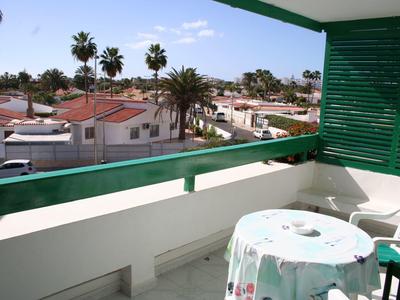 Balkon mit grünem Geländer, Tisch mit Tuch, weiße Häuser und Palmen im Hintergrund unter blauem Himmel.