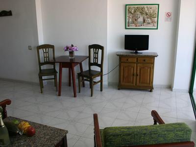 Kleines Wohnzimmer mit weißem Fliesenboden, zwei Stühlen, kleinem Tisch und Fernseher auf Kommode.