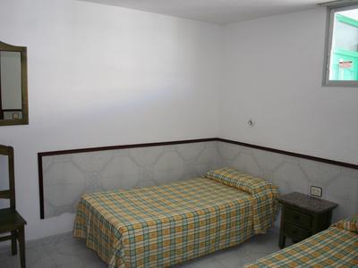 Zimmer mit zwei Betten, karierten Bettdecken, einem Holzstuhl, kleinem Nachttisch und schlichter Wandgestaltung.