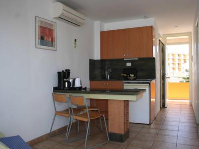 Kleine Küchenzeile mit Holzmöbeln, zwei Stühlen und weißen Wänden, Blick auf Balkon.