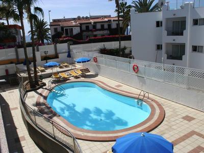 Ein kleiner, ovaler Pool mit blauen Wasser und Liegestühlen umgeben von weißen Gebäuden und Palmen.