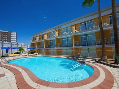 Klare Schwimmbeckenform mit Treppe, umgeben von mehrstöckigem Gebäude und Palmen unter blauem Himmel.