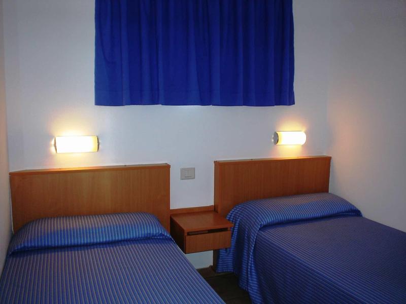 Zwei Einzelbetten mit blauen Decken, Holzkopfenden und wandmontierten Lampen in einem weißen Zimmer.
