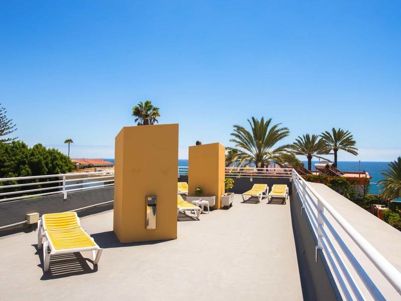 Flacher Balkon mit gelben Liegestühlen, Palmen und Meerblick unter klarem blauem Himmel.