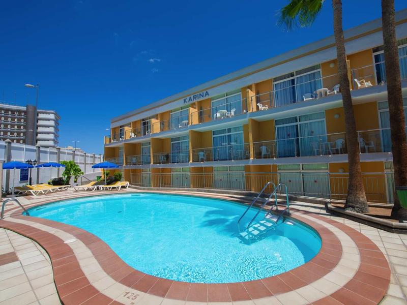 Klare Schwimmbeckenform mit Treppe, umgeben von mehrstöckigem Gebäude und Palmen unter blauem Himmel.