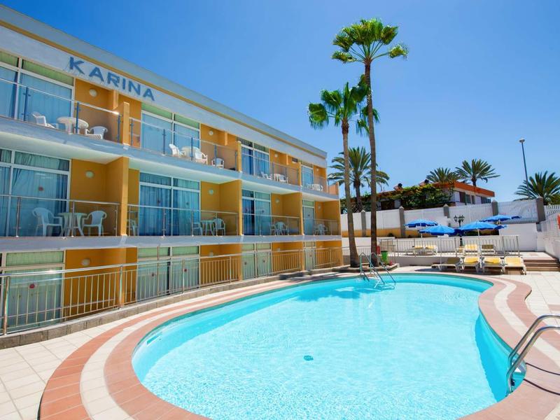 Hellgelbes Hotel mit Balkonen, rundem Pool und Palmen bei strahlend blauem Himmel.