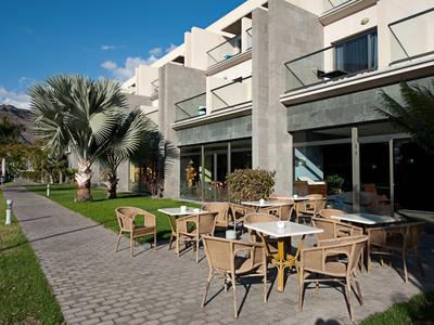 Espace extérieur moderne d'un hôtel avec terrasse, tables, chaises et palmiers sous un ciel bleu.