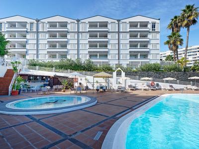 Hotel with outdoor pool, jacuzzi, lounge chairs under blue sky and palm trees.