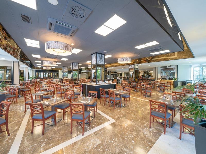 Amplio comedor de hotel con sillas de madera y mesas, suelo de mármol y luz natural.