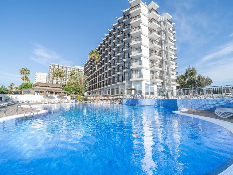 Gran hotel moderno con piscina grande y tumbonas bajo un cielo azul.