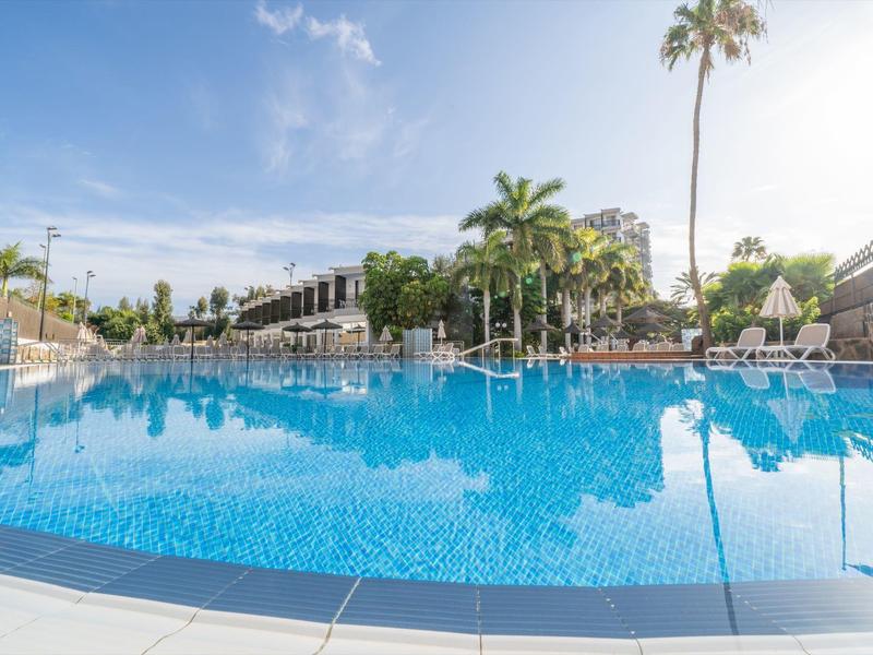 Gran piscina al aire libre con agua azul clara, rodeada de palmeras y edificios del hotel bajo un cielo soleado.