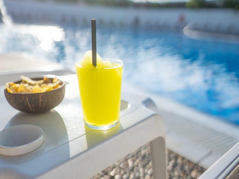 Bebida amarilla y aperitivo en una mesa blanca junto a una piscina con palmeras al fondo.