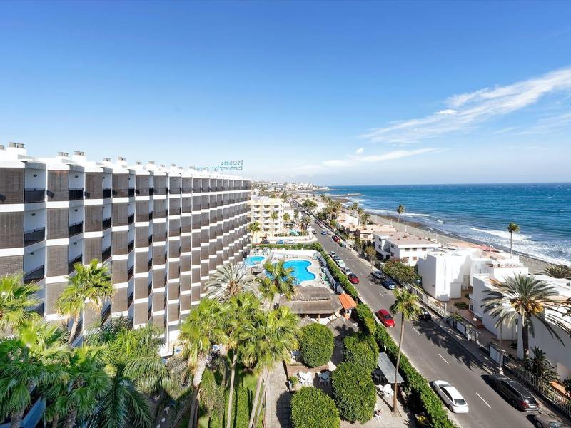 Gran hotel junto al mar con piscina, vista a la calle y palmeras bajo un cielo azul claro.